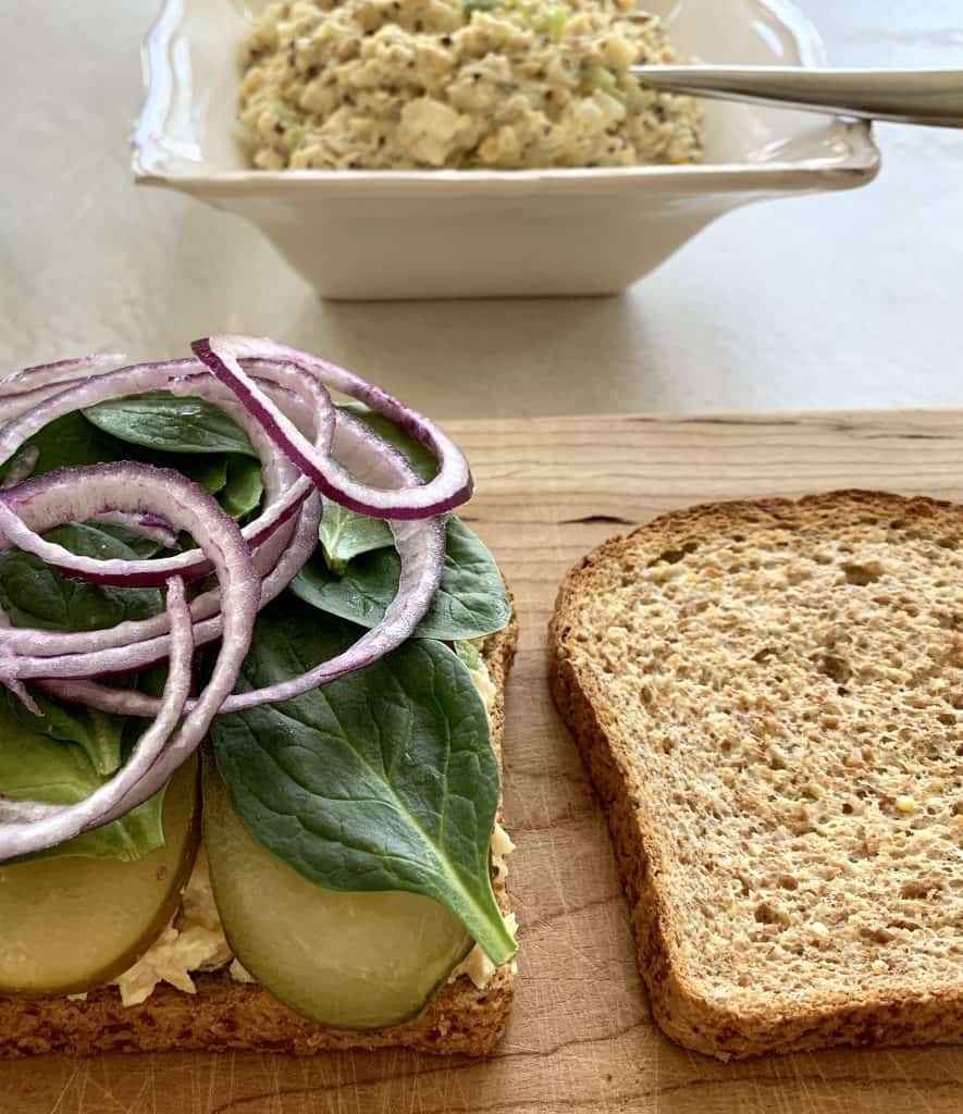 Food on the cutting board consisting of a sandwich made with chickpea salad, greens, red onions, and pickles