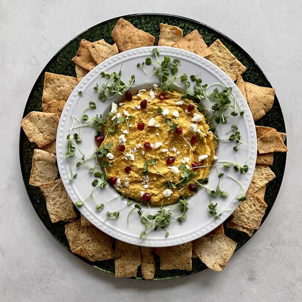 Butternut Squash Hummus & toppings in a white bowl on green plate, surrounded by chips