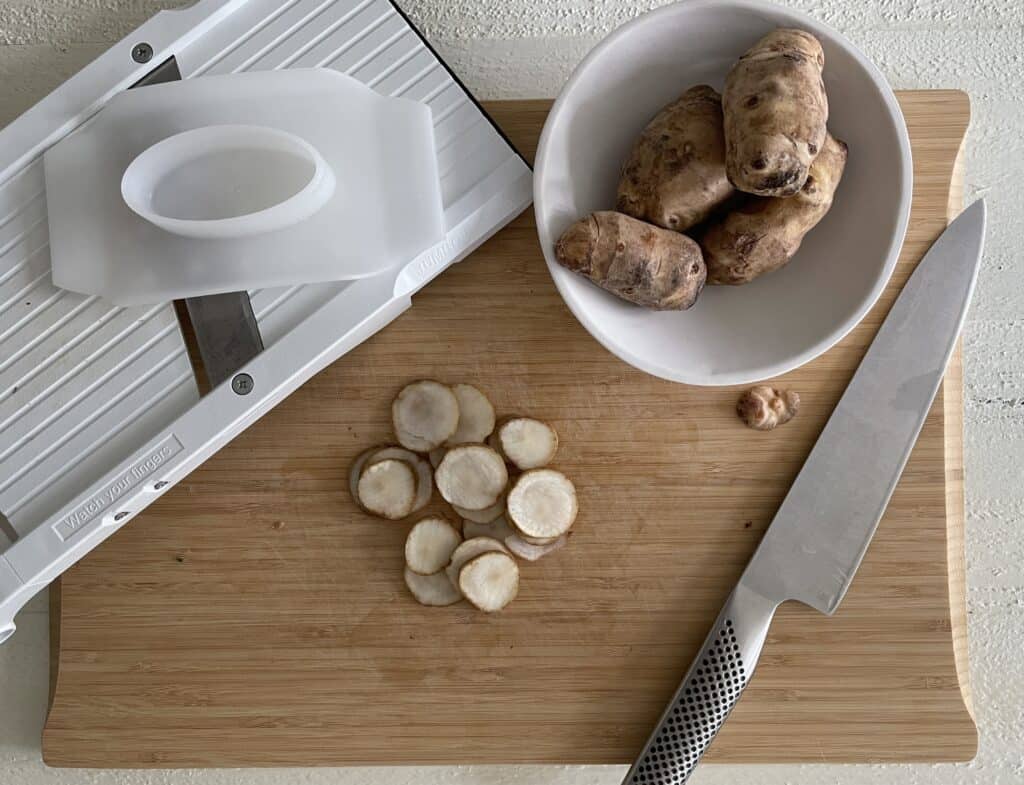 Sunchokes being prepped on a cutting board with a knife and mandoline slicer