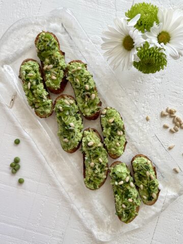 Sweet Pea Crostini on a glass platter with scattered pine nuts, peas and daisies in a bud vase